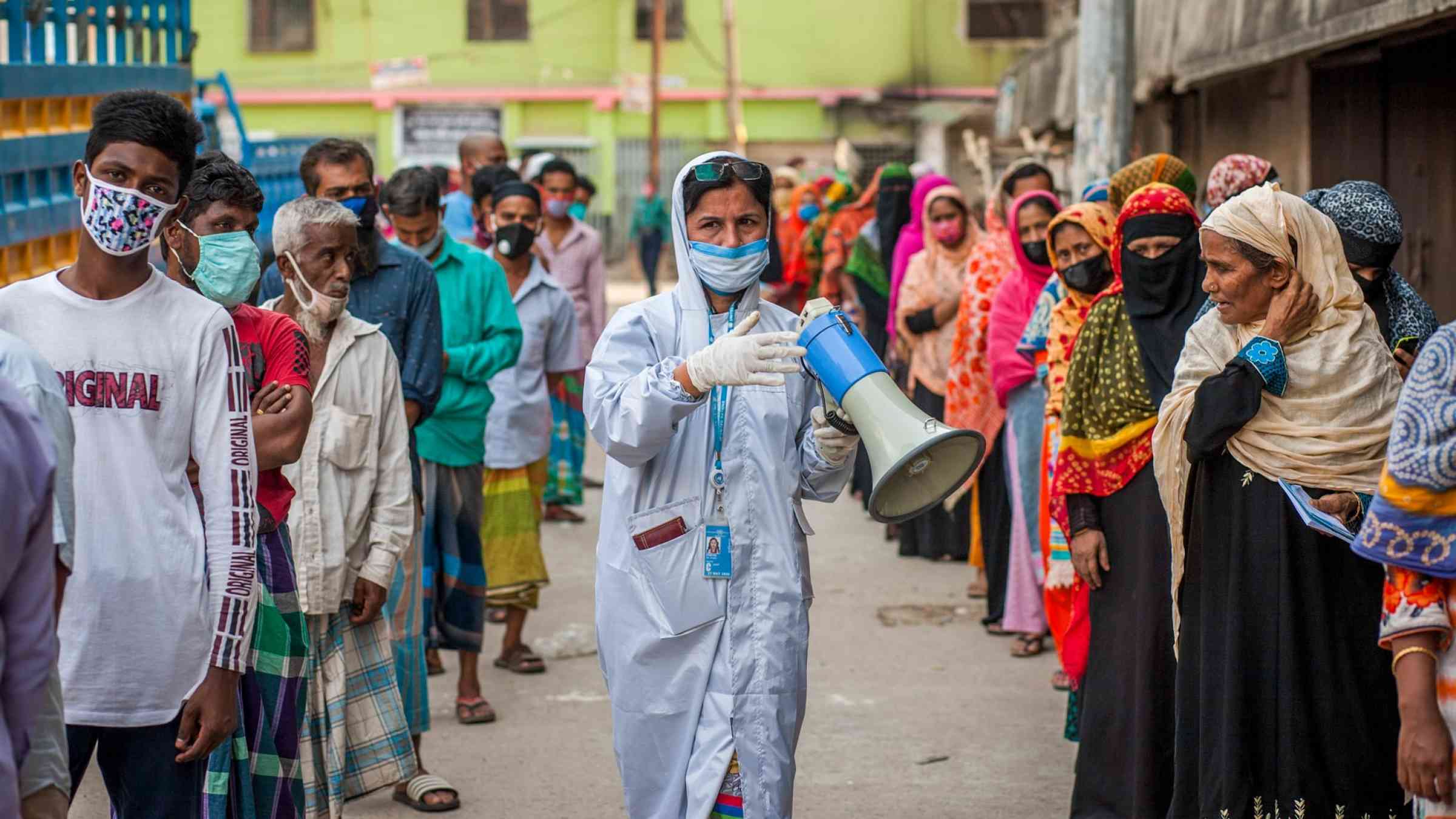 Photo of a woman wearing COVID-19 protective clothing, holding a loudspeaker walking along queuing people in Bangladesh.